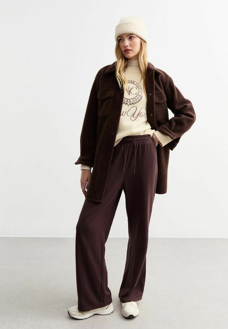 Brauner Wollmantel über einem beigen Sweater mit gedrucktem Text, kombiniert mit dunkelvioletten, weiten Hosen und cremefarbenen Sneakers. Weicher, lässiger Look.