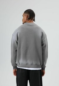 Homme aux cheveux tressés portant un sweat-shirt gris, une chemise blanche en dessous et un jean noir, debout dos à la caméra.