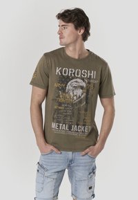 T-shirt verde oliva a maniche corte con stampa grafica raffigurante un'aquila, testo e dettagli consumati. Tessuto in cotone morbido, scollo rotondo.