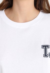 Bílá bavlněná mikina s kulatým výstřihem a námořnickou vyšitou písmenem "T" na levé hrudi. Měkká tkanina, jednoduchý design, bez dalších vzorů.