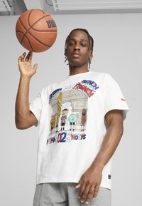 Hvid grafik t-shirt med farverige tegneseriefigurer og tekst, med en løs pasform. Modellen holder en basketball og har grå shorts på.