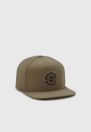 Brązowa czapka snapback z okrągłą naszywką z napisem „"Brixton Supply Trade Mark"“ na przednim panelu, pokazana na jednolitym jasnym tle.