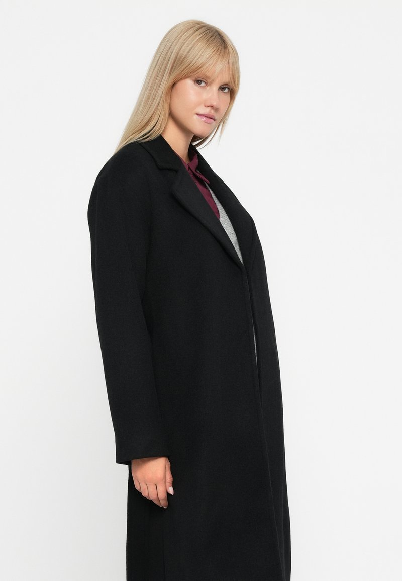 Lång svart kappa med skräddarsydd design, slät krage och långa ärmar. Slät textur, medellång, med en framknäppning.