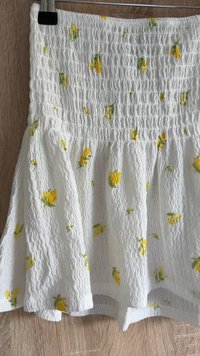 Fehér, textúrált szoknya apró sárga virágmintákkal. Smockolt derékrésszel és bővülő aljjal rendelkezik a dúsabb hatás érdekében.