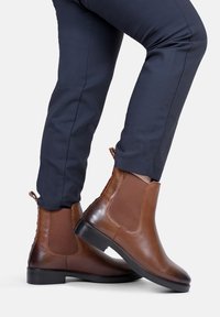 Brązowe skórzane botki Chelsea z elastycznymi panelami bocznymi i teksturowaną podeszwą, charakteryzujące się gładkim wykończeniem i niskim obcasem blokowym. Noszone z granatowymi spodniami.