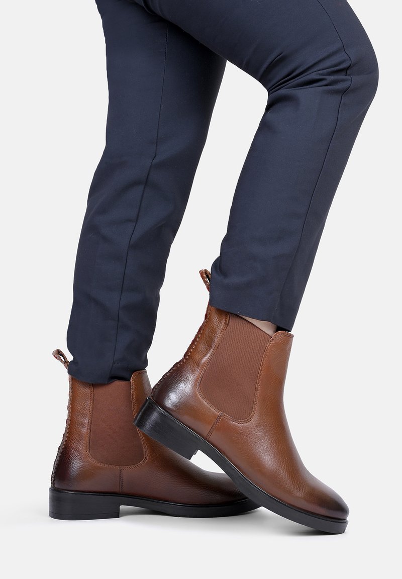 Brązowe skórzane botki Chelsea z elastycznymi panelami bocznymi i teksturowaną podeszwą, charakteryzujące się gładkim wykończeniem i niskim obcasem blokowym. Noszone z granatowymi spodniami.