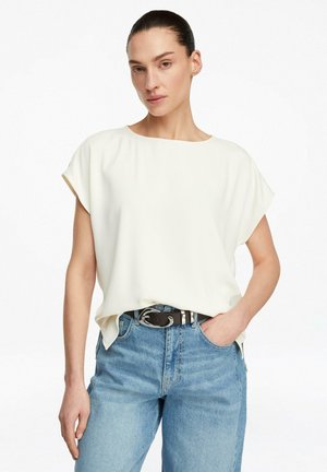 Kobieta o ciemnych włosach związanych z tyłu, ubrana w luźną białą bluzkę z krótkim rękawem, wpuszczoną w jasnoniebieskie dżinsy z czarnym paskiem, stojąca na tle białego tła.