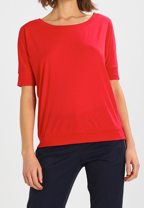 Femme portant un haut rouge vif à manches courtes avec un col rond, associée à un pantalon bleu marine foncé sur un fond uni.
