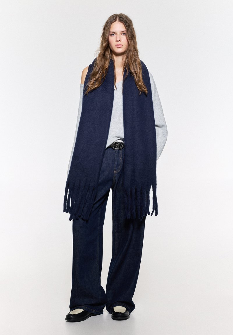 Mørkeblå frynset tørklæde lavet af blødt stof, lang og rektangulær form, draperet omkring skuldrene, parret med en lysegrå sweater og jeans.