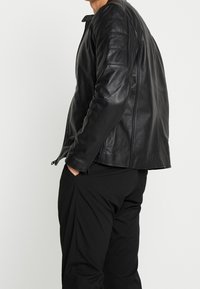 Giubbotto di pelle nera con dettagli trapuntati sulle spalle e zip laterali. Indossato sopra pantaloni neri, evidenziando una vestibilità slanciata e una texture liscia.