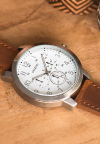 Round stainless steel watch with white dial, black numerals, date display, and brown leather strap. Features include sub-dials and metal accents.