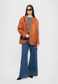Donna che indossa una giacca di pelle marrone, una maglietta a righe, jeans a gamba larga, scarpe nere a punta, occhiali da sole e tiene una piccola borsa a spalla nera.