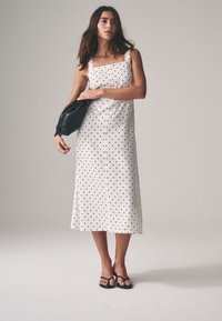 White sleeveless dress with black polka dots, square neckline, and flowing hem. Paired with a black clutch and black flip-flops.