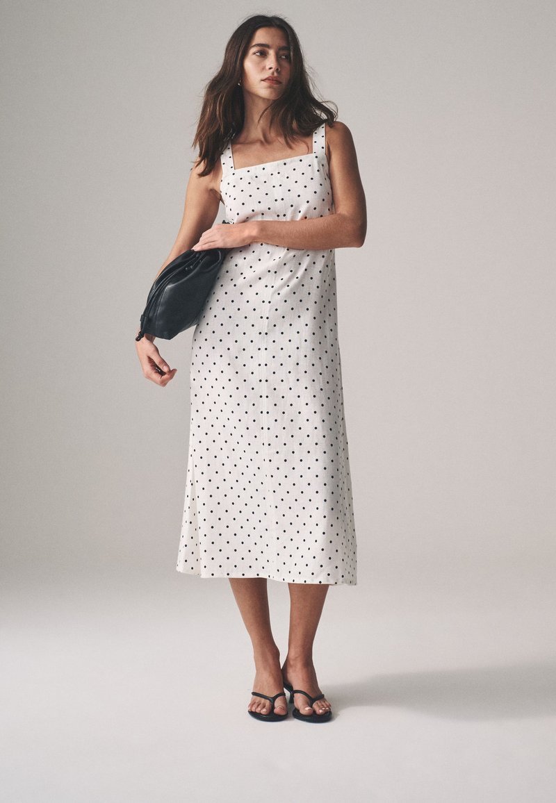 White sleeveless dress with black polka dots, square neckline, and flowing hem. Paired with a black clutch and black flip-flops.