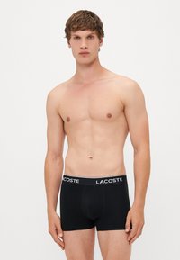 Des boxers noirs en coton avec une taille élastique arborant le logo "LACOSTE" en blanc. Texture lisse, design ajusté.