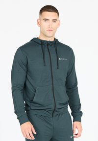 Mörkgrön zip-up hoodie i mjukt tyg, utrustad med en huva, justerbara snörr, framfickor och en subtil logotyp på bröstet.