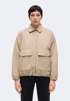 Personne aux cheveux courts et foncés porte une veste beige avec deux grandes poches avant et un pantalon noir, debout devant un fond clair uni.