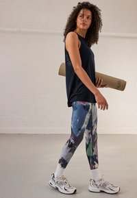 Top nero senza maniche, leggings con fantasia astratta colorata, sneakers bianche con dettagli grigi. Tiene in mano un tappetino da yoga arrotolato con custodia marrone.