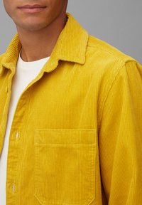 Camisa de pana amarilla con superficie texturizada, que presenta un cuello extendido y un bolsillo en el pecho, usada sobre una camiseta blanca.