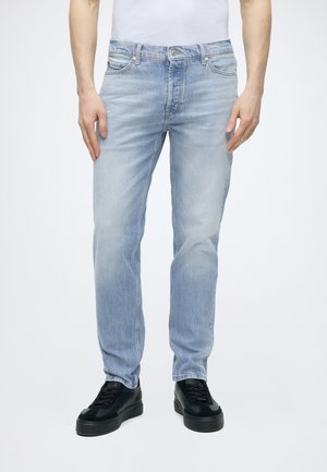 Man draagt lichtblauwe jeans met rechte pijpen en zwarte sneakers, staat voor een witte achtergrond, handen ontspannen langs het lichaam.