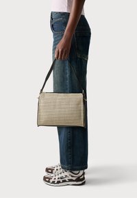 Borsa rettangolare con un motivo a griglia testurizzato beige e nero, dotata di una tracolla nera. Abbinata a un paio di jeans blu larghi e scarpe da ginnastica.