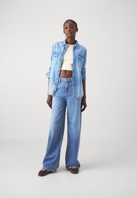 Chemise en denim bleu clair avec col pointu, deux poches avant et fermeture à boutons, associée à un jean bleu clair à jambes larges et des chaussures foncées.