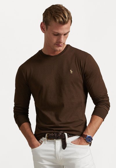 Brown long-sleeve T-shirt with a round neckline and a small gold logo on the chest, paired with white pants and a woven brown belt.