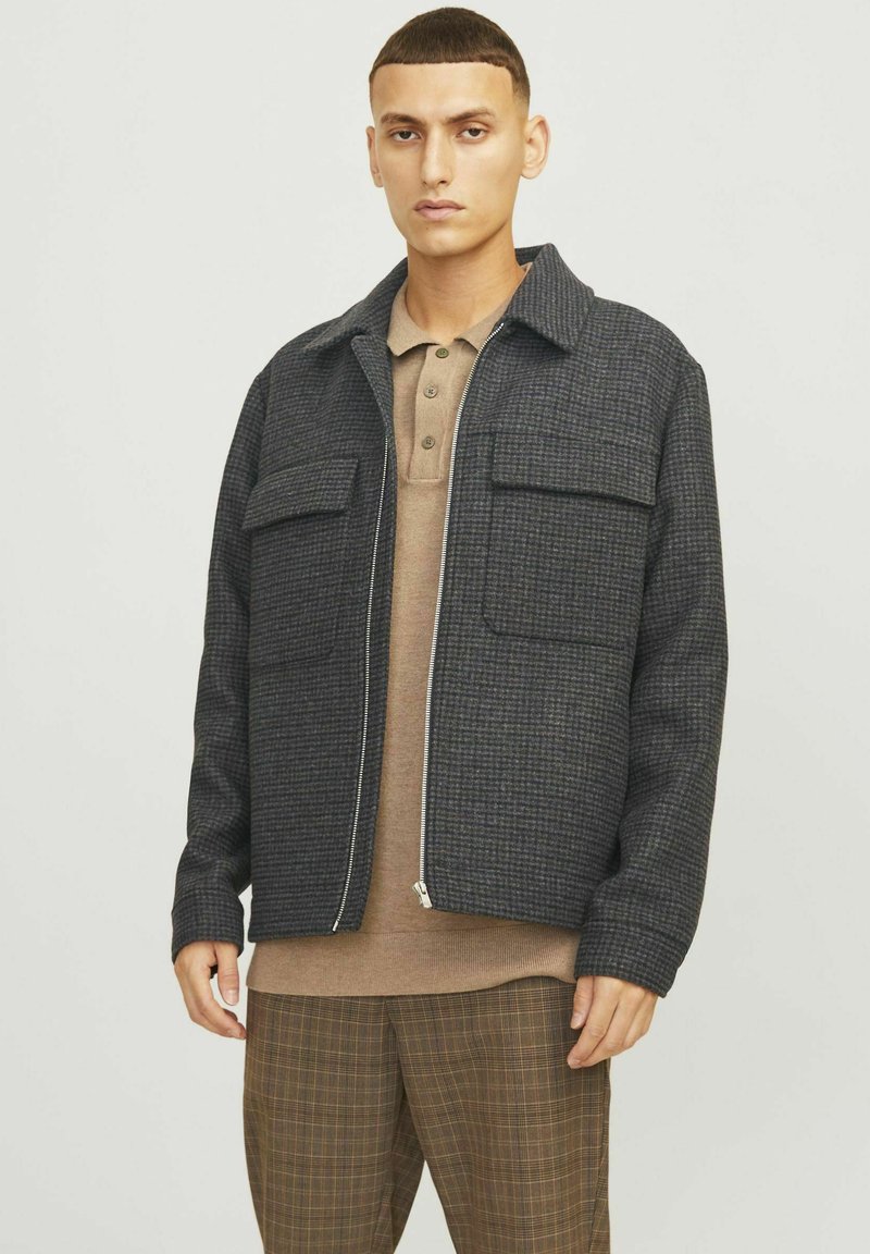 Veste à carreaux gris avec une fermeture éclair à l'avant, deux poches poitrine et un col, portée sur un polo marron, assortie à un pantalon à carreaux marron.