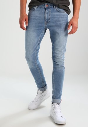 Mann trägt hellblaue enge Jeans mit Umschlägen und weiße High-Top-Sneakers, steht vor einem schlichten Hintergrund.