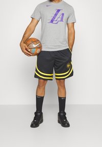 Šedé tričko Nike s fialovým grafickým motivem "LA", černé šortky se žlutými akcenty, černé ponožky a černé tenisky. V jedné ruce drží basketbal.