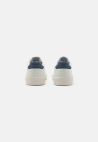 Hvite joggesko med en marineblå hælkappe, teksturert lærøverkledning og off-white gummisåle. Minimalistisk design med sømdetaljer.