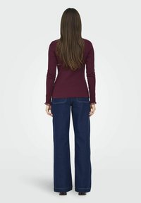Maglia a maniche lunghe color bordeaux con design aderente abbinata a jeans a gamba larga blu con tasche e cuciture a contrasto. Vista da dietro.
