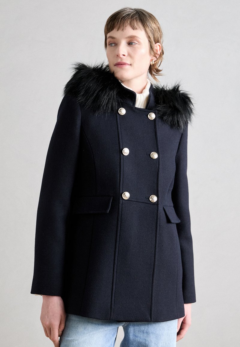Tamsiai mėlynas dvigubo užsegimo paltas su dirbtiniu kailiu, sidabrinėmis sagomis ir priekinėmis kišenėmis. Tekstūruotas audinys ir prigludęs dizainas.