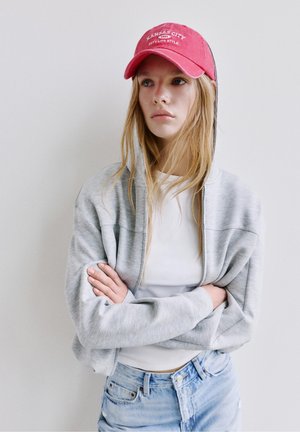 Mladi žena s dugom plavom kosom koja nosi crvenu kapu Kansas Cityja, svijetlosivu zip-up hoodicu, bijelu majicu i plave traperice, stoji sa sklopljenim rukama.