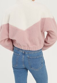 Kurz geschnittene Fleecejacke in Pink und Weiß mit Chevron-Muster, ausgestattet mit hohem Kragen und elastischem Bund. Kombiniert mit blauen Jeans.