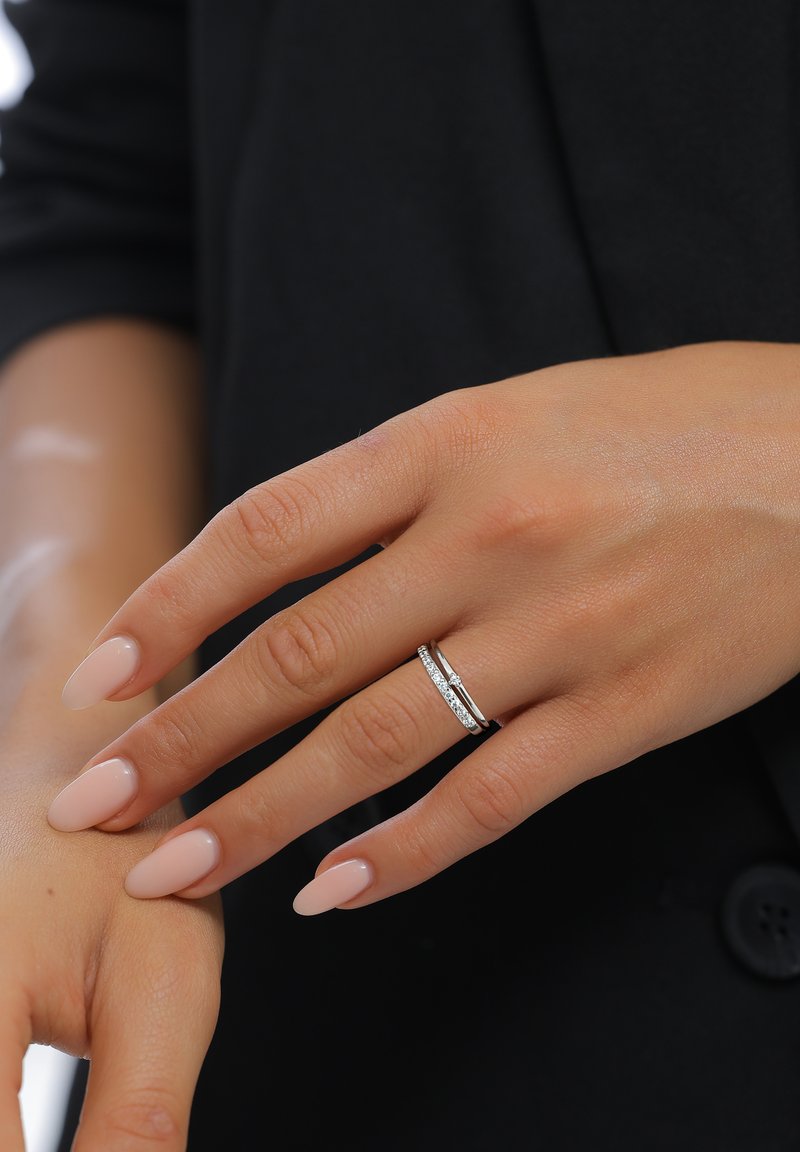 Bague en argent avec une fine bande, ornée d'une rangée de petites pierres claires. La main a des ongles soigneusement manucurés de couleur rose pâle. Le fond est noir.