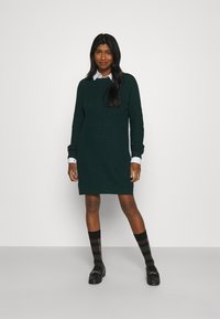 Vestido de malha verde escuro com gola redonda e mangas longas, combinado com uma camisa branca de colarinho, sapatos pretos e meias até ao joelho em padrão xadrez.