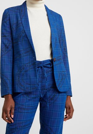 Blauw geruit pak met zwarte schrifttekst, bestaande uit een enkelknopsblazer en een broek met trekkoord, gedragen over een witte coltrui.