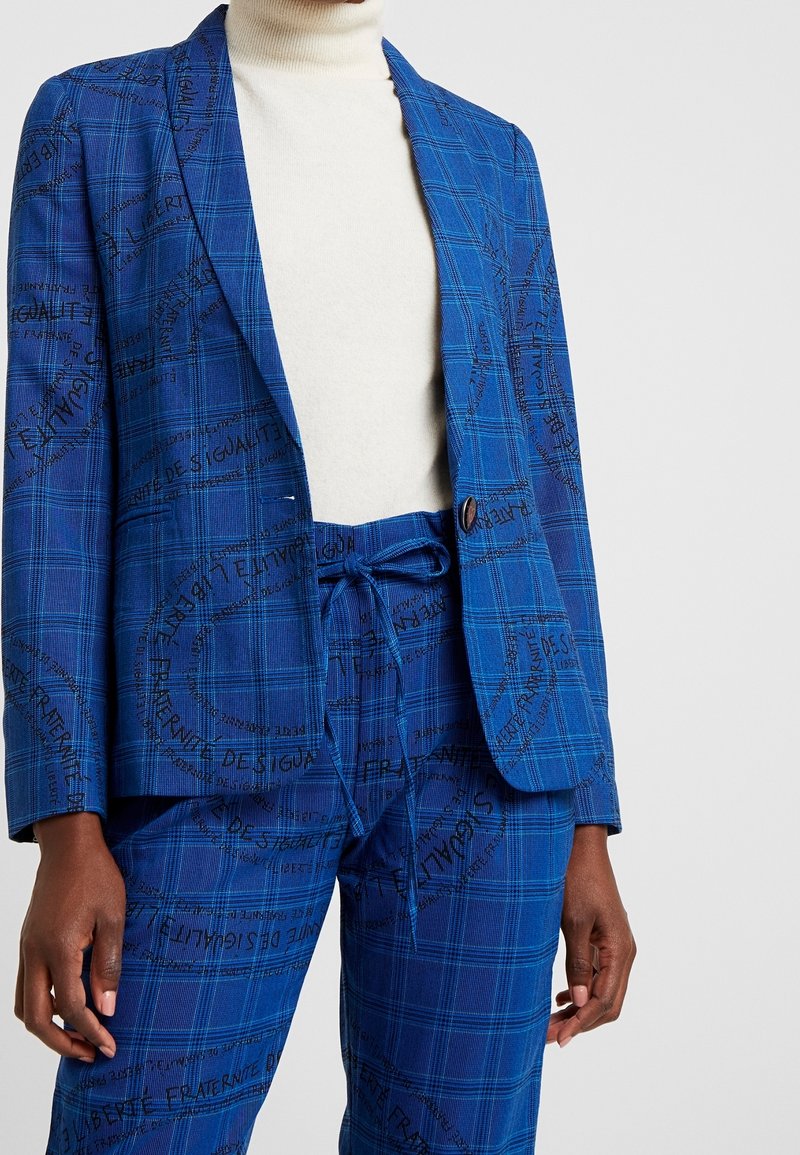 Costume à carreaux bleus avec texte manuscrit noir, comprenant un blazer à un bouton et un pantalon avec cordon, porté sur un pull à col roulé blanc.