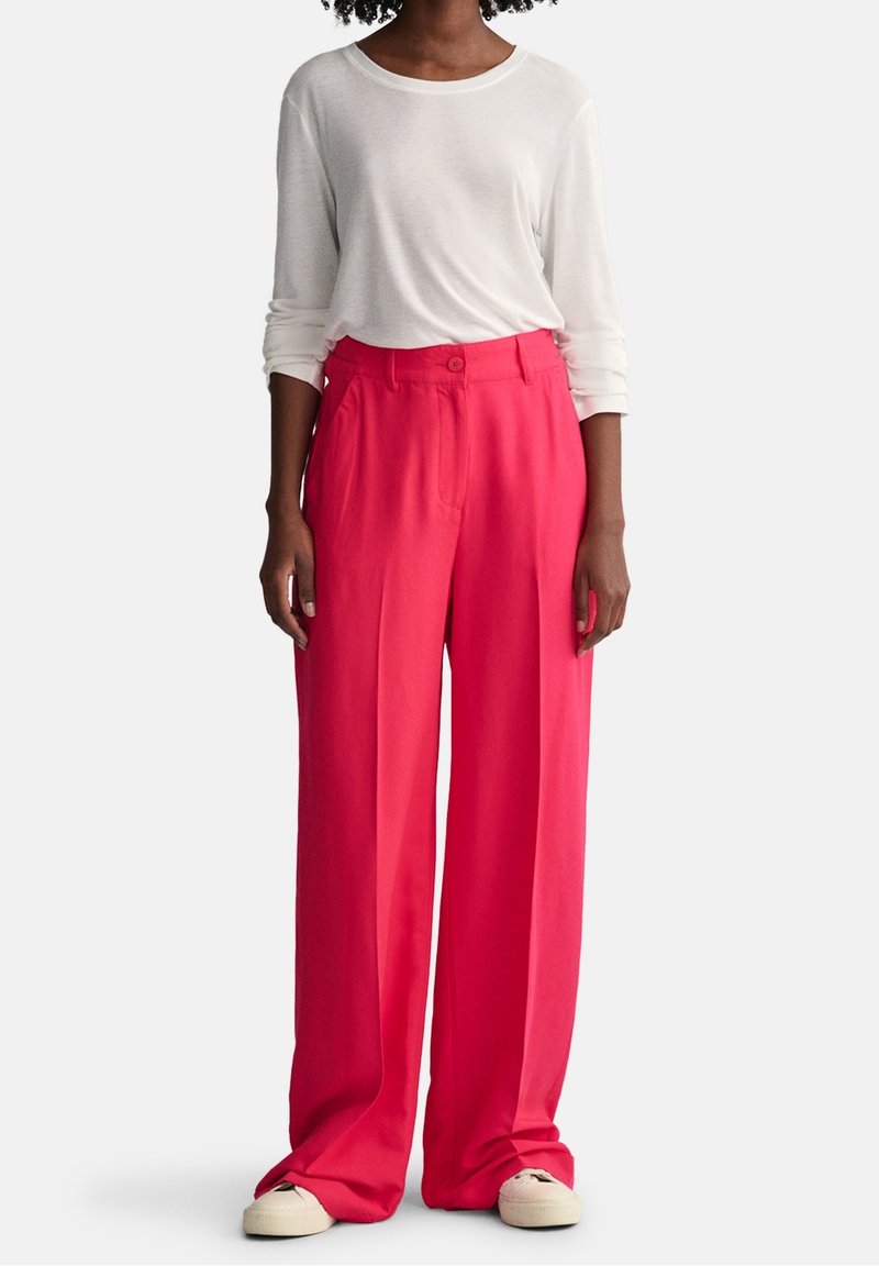 Femme portant un pantalon large ample rose vif, un haut blanc à manches longues et des baskets blanches, debout devant un fond clair.