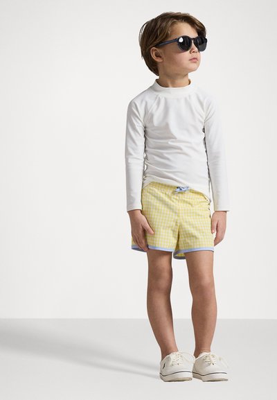 Camisa blanca de manga larga, pantalones cortos de baño a cuadros amarillos con ribete azul, y zapatillas blancas. Niño con gafas de sol negras, en pose de pie.