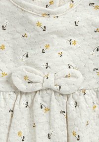 Robe beige clair avec des imprimés floraux jaunes et blancs, dotée d'un tissu texturé, d'un col rond et d'un accent en nœud à la taille.