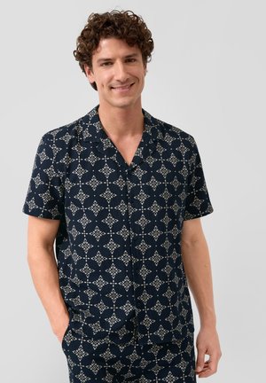 Man met krullend haar die een marineblauw overhemd met korte mouwen en witte geometrische patronen draagt, lacht met zijn handen in zijn zakken.