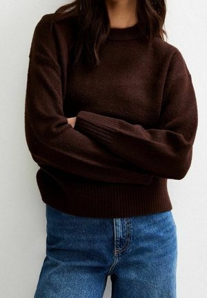 Kvinde iført en mørkebrun langærmet sweater og blå højtaljede jeans, stående med armene krydsede.