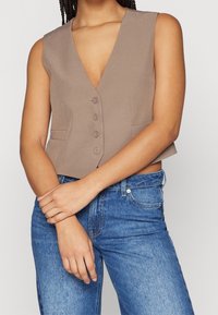Ljusbrun, ärmlös väst med djup V-hals och fem knäppningar; har två sidofickor och en kort design. Matchad med blå jeans.