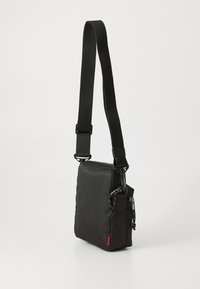 Compact black textured crossbody bag with adjustable strap, zip closure, and small red tag at bottom corner.