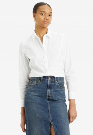 Mujer con el cabello peinado hacia atrás, vistiendo una camisa blanca abotonada y una falda vaquera de talle alto con una abertura frontal, posando contra un fondo liso.