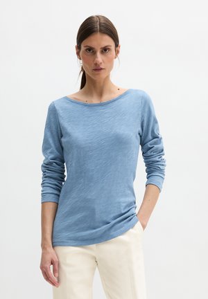 Vrouw met donker haar in een staart, gekleed in een lichtblauw shirt met lange mouwen en gebroken witte broek, staand tegen een effen lichte achtergrond.