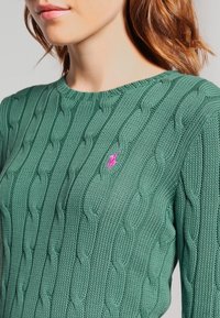 Grön stickad tröja med flätmönster, med rund halsringning och en liten rosa logotypdetalj på bröstet.