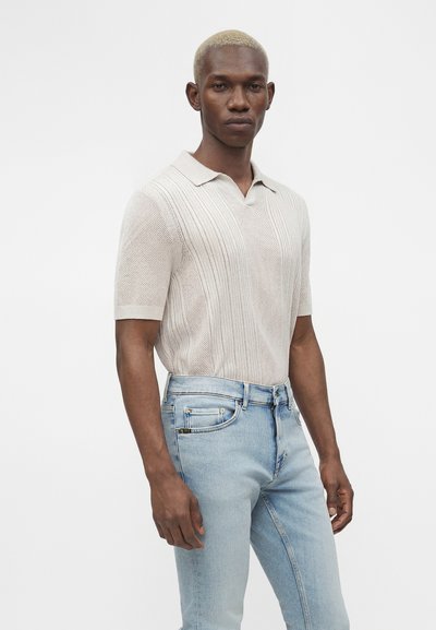 Άνδρας με κοντά ξανθά μαλλιά, φοράει ανοιχτό μπεζ κοντομάνικο πλεκτό πόλο και ανοιχτό μπλε τζιν ψηλόμεσο, στέκεται πλαγίως.